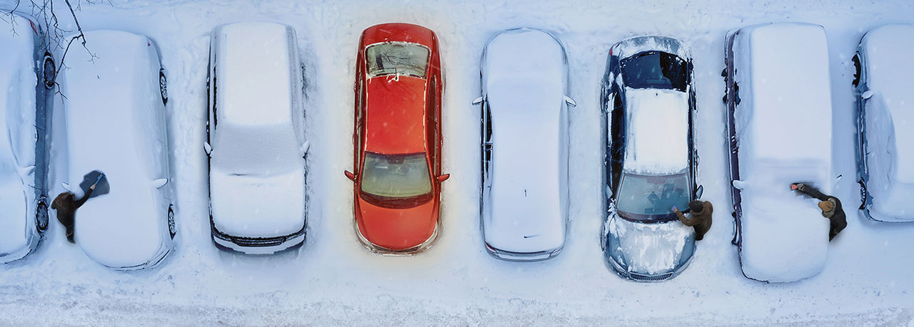 Verschneite Pkw auf einem Parkplatz, ein roter, schneefreier Wagen und räumende Personen; Visualisierung für Presse zu Standheizungen – Webasto als Systempartner.