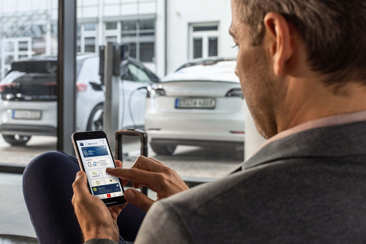 Business user managing EV charge data via Webasto charging app with vehicles and charger in background, showcasing Webasto as vehicle electrification system partner.